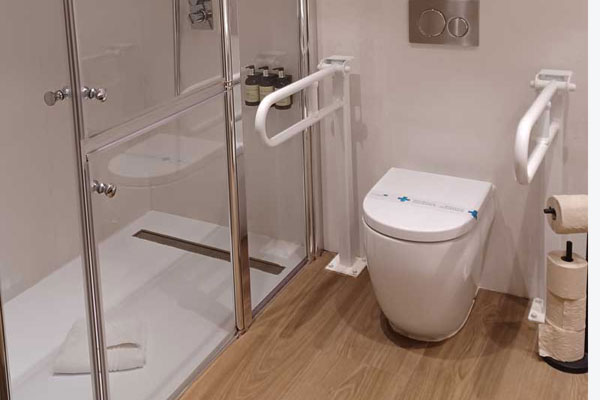 Barras de apoyo abatibles a ambos lados del inodoro y plato de ducha con mampara del aseo de la habitación adaptada. 