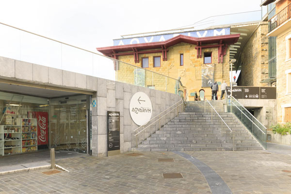 Exterior del Aquarium, entrada señalizada con un cartel blanco y un totem informativo negro. Leve rampa de acceso y puertas automáticas de cristal.