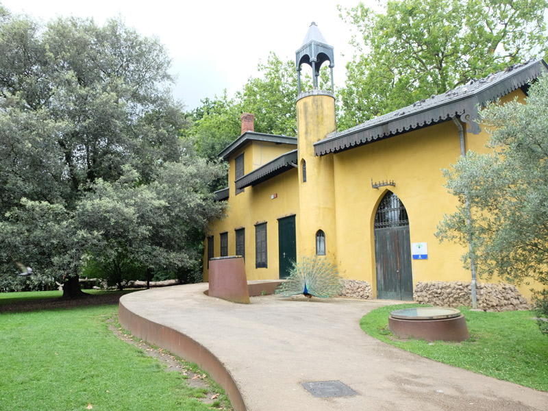 Edificio de las cocinas y la capilla del parque. Camino asfaltado hasta la puerta del establecimiento con fachada amarilla, un pavo real posado junto a la entrada.