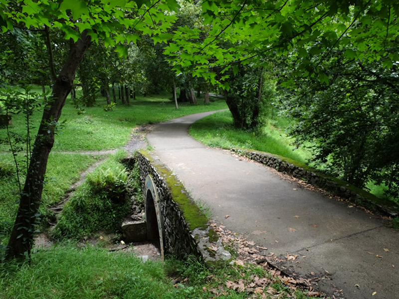 Section of the asphalt path of the Cristina-Enea park that passes over a stone bridge, trees and green areas on both sides.